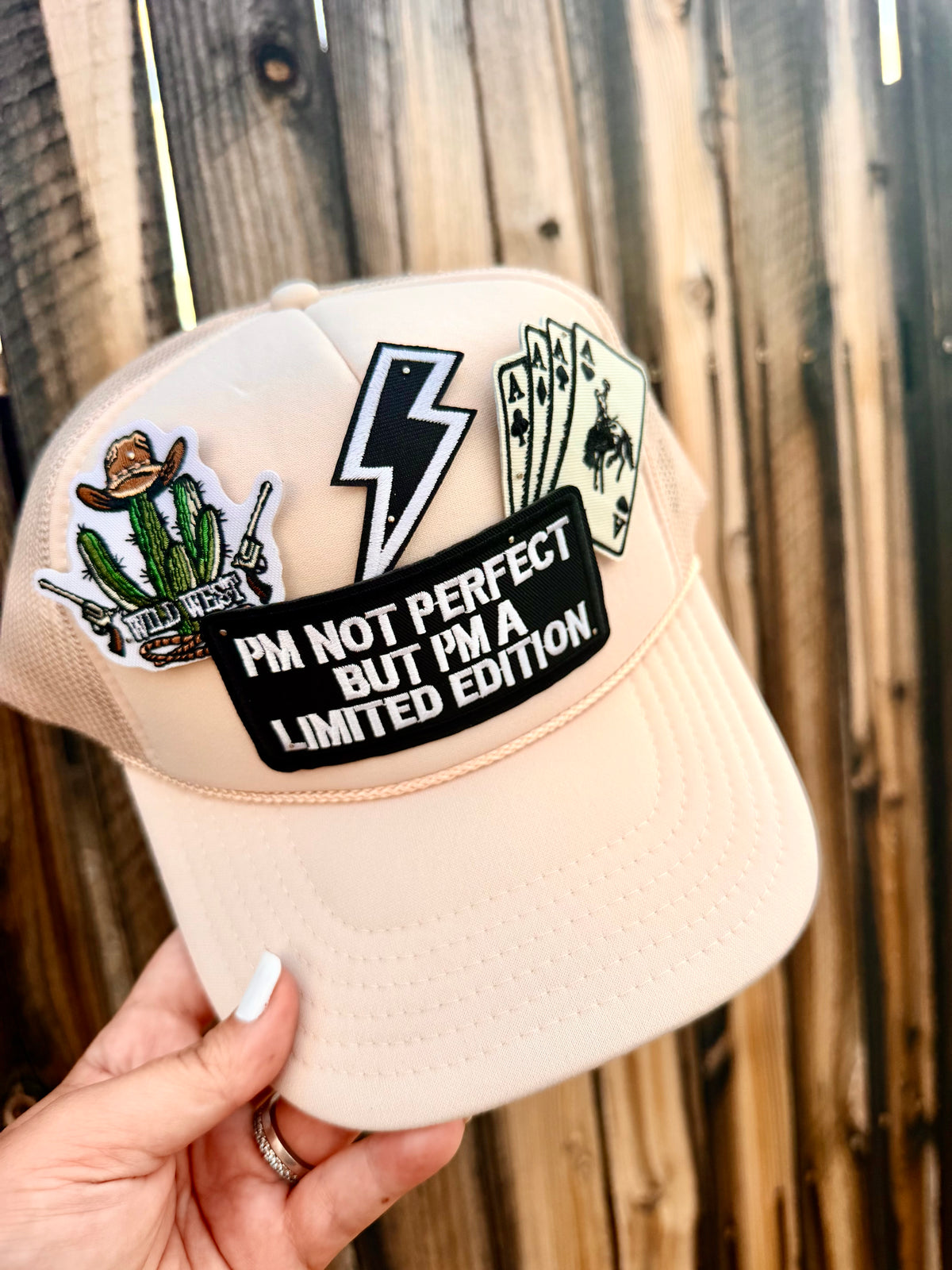 Beige cap with various patches including a cactus, lightning bolt, and text, held against a wooden background.